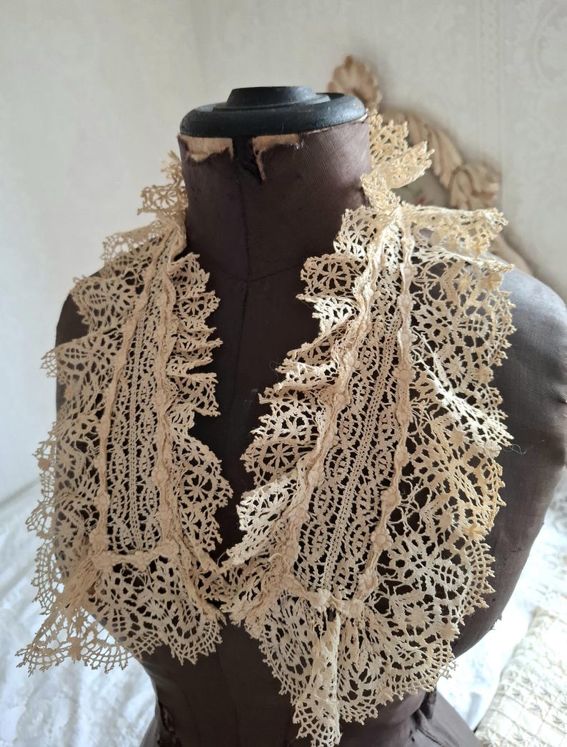 May include: An antique cream-colored lace collar with intricate floral and geometric patterns. The collar is draped over a brown mannequin, showcasing its delicate, openwork design and ruffled edges. The vintage textile adds a touch of elegance.