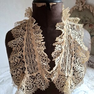 May include: An antique cream-colored lace collar with intricate floral and geometric patterns. The collar is displayed on a vintage brown dress form. The lace has a delicate, openwork design with scalloped edges, showcasing a vintage aesthetic.