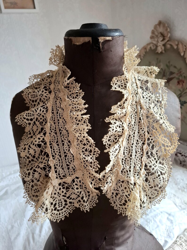 May include: An antique cream-colored lace collar with intricate floral and geometric patterns. The collar is displayed on a vintage brown dress form. The lace has a delicate, openwork design with scalloped edges, showcasing a vintage aesthetic.