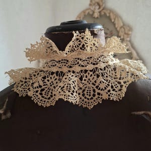 May include: An antique cream-colored lace collar, featuring intricate openwork patterns. The collar is displayed on a dark brown mannequin, showcasing its delicate design and vintage aesthetic. The lace is likely cotton or linen.