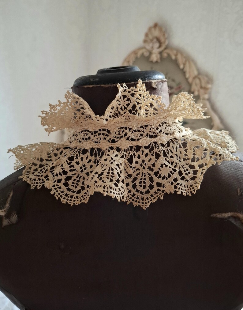 May include: An antique cream-colored lace collar, featuring intricate openwork patterns. The collar is displayed on a dark brown mannequin, showcasing its delicate design and vintage aesthetic. The lace is likely cotton or linen.