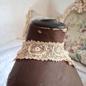 May include: A cream-colored, antique lace collar with a delicate floral pattern. The collar is displayed on a brown mannequin, showcasing its intricate design and vintage aesthetic. The lace is finely detailed.