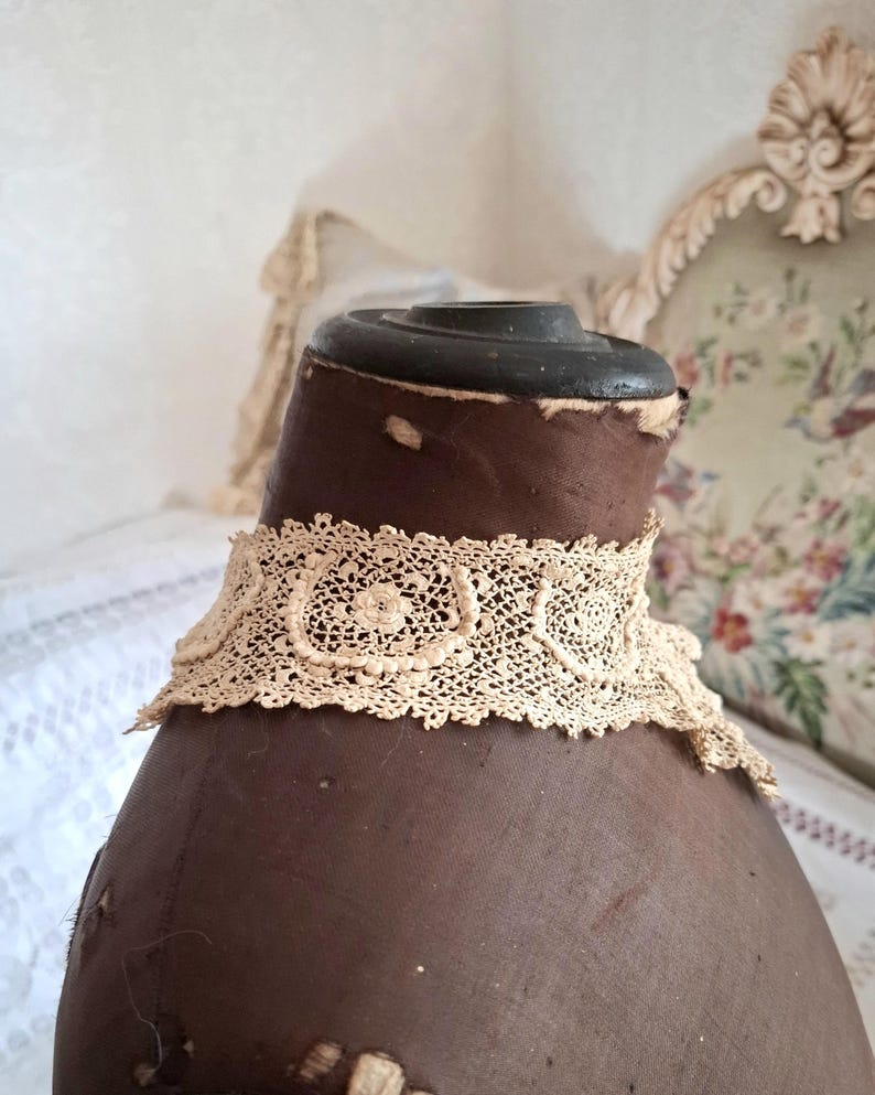 May include: A cream-colored, antique lace collar with a delicate floral pattern. The collar is displayed on a brown mannequin, showcasing its intricate design and vintage aesthetic. The lace is finely detailed.