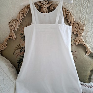 May include: A plain white sleeveless slip dress featuring a square neckline and wide shoulder straps. The garment is constructed from a solid white fabric and includes subtle horizontal stitched detailing across the upper chest area.