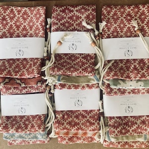Könnte beinhalten: Sechs rechteckige Stoffbeutel mit einem sich wiederholenden Blumenmuster in Rot- und Weißtönen. Jeder Beutel ist mit einer weißen Papierbanderole mit dem Aufdruck Skye & Loom versehen und hat weiße Kordelzüge mit kleinen Holzperlen.