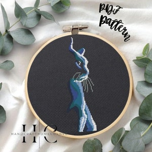 May include: A cross-stitch pattern of a blue and green cat on a black background. The pattern is in a wooden embroidery hoop. The text "PDF Pattern" is written in white on a white background.