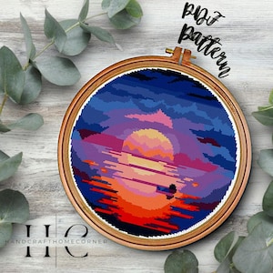 May include: A cross stitch pattern of a sunset over a body of water. The sunset is a vibrant orange, yellow, and pink. The water is a deep blue with a reflection of the sunset. The pattern is in a wooden embroidery hoop.