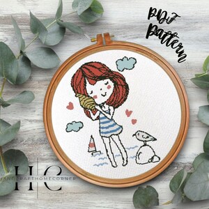 Pode incluir: Um padrão de ponto cruz com uma menina de cabelo ruivo segurando uma concha no ouvido. Ela está vestindo um maiô listrado azul e branco e está em pé em uma praia com uma gaivota e um veleiro ao fundo. O padrão é emoldurado em um aro de madeira.