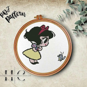Puede incluir: Patrón de punto de cruz con una ilustración de dibujos animados de Blancanieves con un lazo rojo en el pelo, vistiendo un vestido amarillo y sosteniendo una manzana roja. Un pequeño pájaro está en el fondo. El patrón está en un aro de bordado de madera.