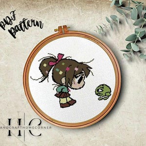 May include: Cross stitch pattern featuring a girl with brown hair and a pink bow, holding a green frog. The pattern is displayed in a wooden embroidery hoop.