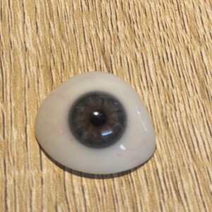 Könnte beinhalten: Ein Augenprothese mit einer dunkelbraunen Iris und einer schwarzen Pupille, eingebettet in eine weiße Sklera, die feine, realistische rote Gefäßdetails aufweist. Die Oberfläche ist glatt und abgerundet und ahmt die natürliche Form nach.
