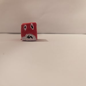 May include: A small red cube with a white face and black markings that resemble a face.