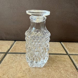 May include: A clear, vintage-style glass bottle with a decorative stopper. The bottle has a diamond-patterned design on the lower portion and a flared top. The stopper is a flat, circular shape. The bottle is approximately 6 inches tall.