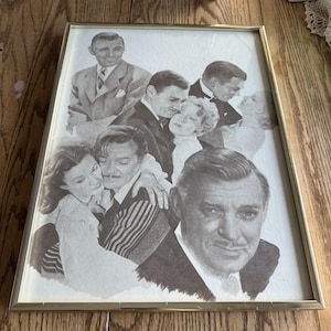 May include: A framed black and white print featuring multiple portraits of actors in vintage attire. The print has a gold-colored frame and is displayed on a wooden surface. The image appears to be a collage of classic Hollywood stars.
