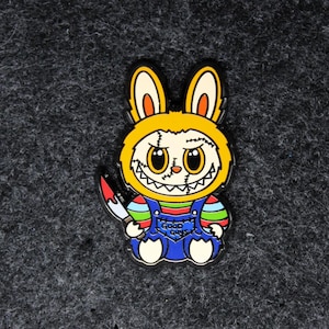 Può includere: Una spilla smaltata colorata raffigurante un personaggio coniglio con cappuccio giallo e tuta blu. Il coniglio tiene in mano un coltello insanguinato e ha la faccia cucita. Il testo "GOOD GUYS" è sulla tuta.