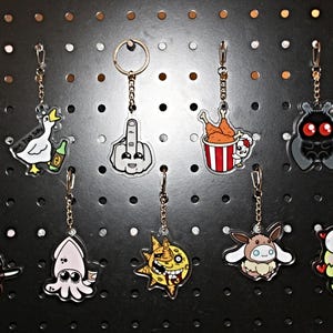 Op de afbeelding: Een verzameling novelty sleutelhangers met verschillende cartoon- en popcultuurontwerpen. De sleutelhangers tonen personages als Yoshi, een eend met bier en een middelvinger-emoji. Elke sleutelhanger is bevestigd aan een metalen ketting en sluiting.