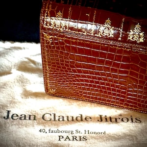May include: A brown leather handbag with a crocodile skin texture and gold accents. The bag is resting on a white cloth with the text "Jean Claude Jitrois, 40, faubourg St. Honoré PARIS". The bag has a rectangular shape.
