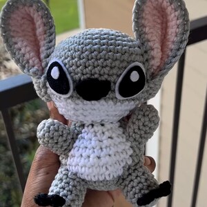 Puede incluir: Un juguete de peluche gris tejido a crochet con grandes ojos negros y una barriga blanca. El juguete tiene orejas grandes y una nariz negra cosida.
