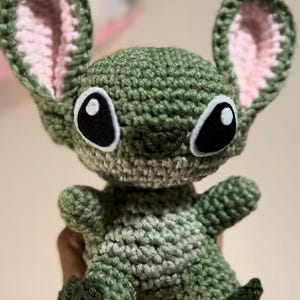 Puede incluir: Juguete de peluche verde tejido a crochet con orejas rosas y ojos negros. El juguete tiene forma de un extraterrestre de dibujos animados con una amplia sonrisa.