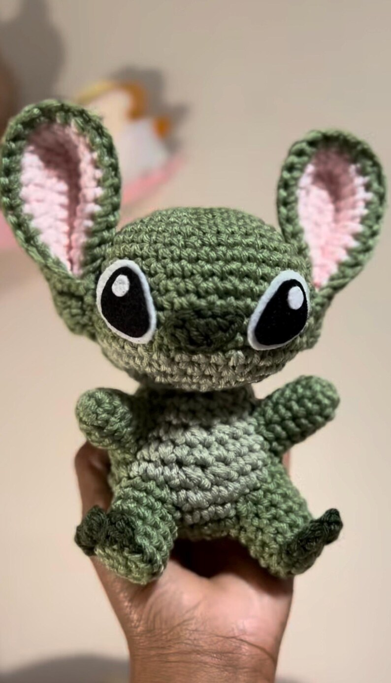 Puede incluir: Juguete de peluche verde tejido a crochet con orejas rosas y ojos negros. El juguete tiene forma de un extraterrestre de dibujos animados con una amplia sonrisa.