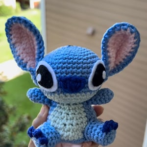 Puede incluir: Un juguete de peluche Stitch de crochet azul y rosa con ojos negros y un contorno blanco. El juguete est&aacute; siendo sostenido en una mano.