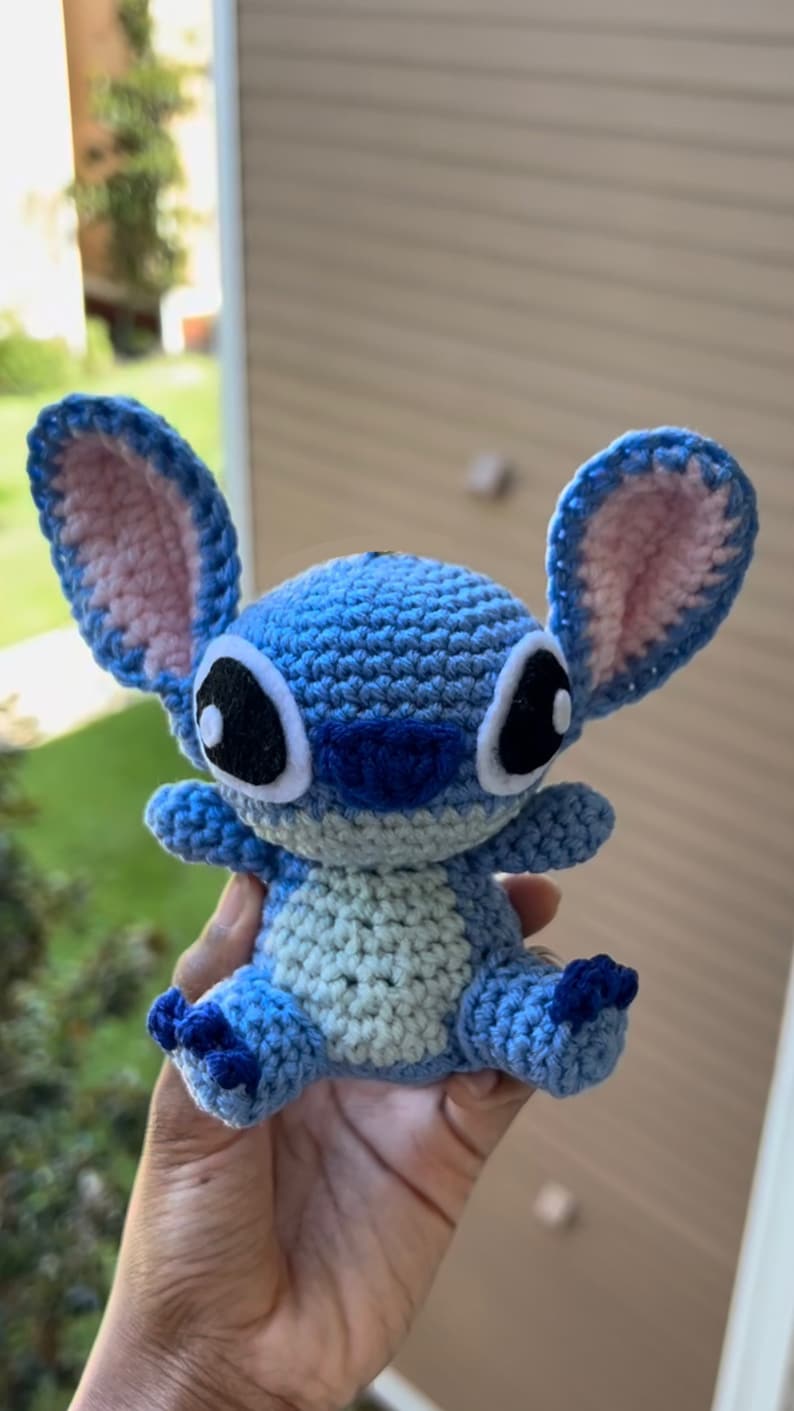 Puede incluir: Un juguete de peluche Stitch de crochet azul y rosa con ojos negros y un contorno blanco. El juguete est&aacute; siendo sostenido en una mano.