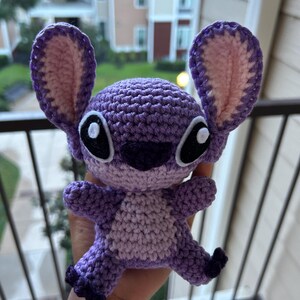 Puede incluir: Juguete de peluche Stitch de color morado tejido a crochet con orejas rosas y ojos negros.
