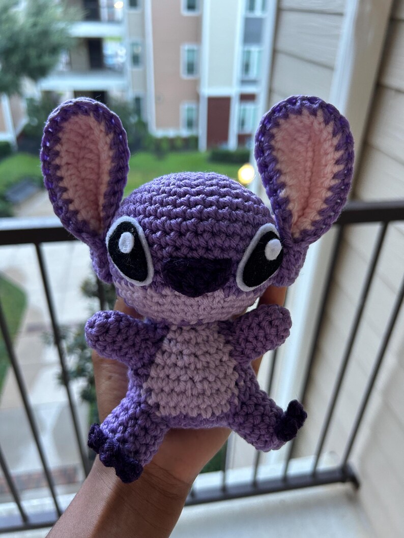 Puede incluir: Juguete de peluche Stitch de color morado tejido a crochet con orejas rosas y ojos negros.