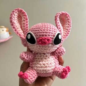 Puede incluir: Un juguete de peluche Stitch de crochet rosa con ojos negros y una nariz rosa. El juguete est&aacute; sentado en una mano.
