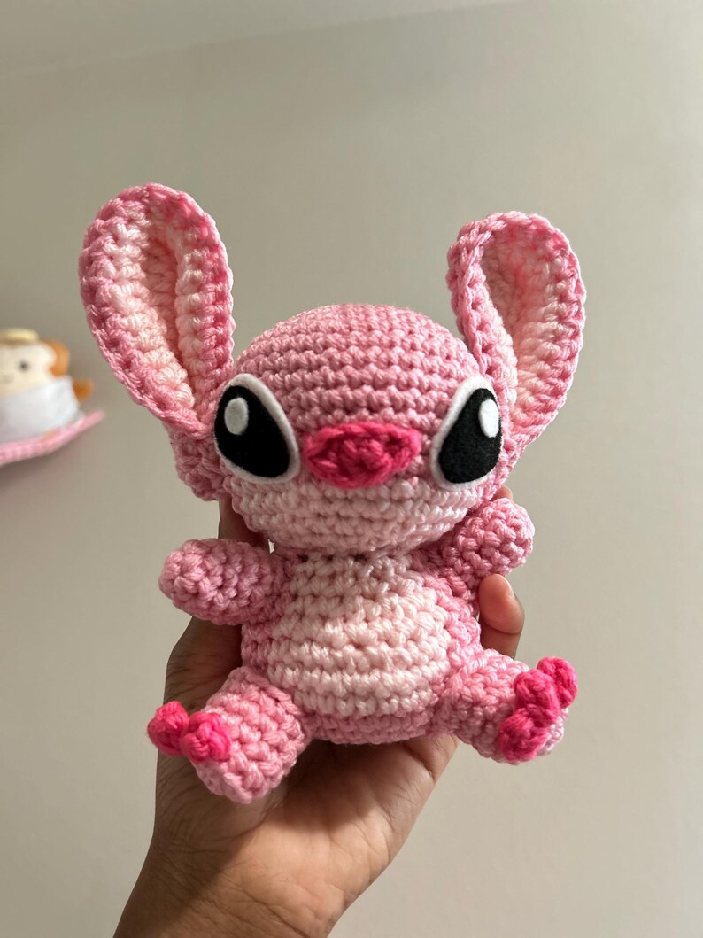 Puede incluir: Un juguete de peluche Stitch de crochet rosa con ojos negros y una nariz rosa. El juguete est&aacute; sentado en una mano.