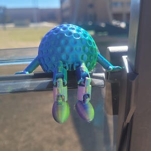 Puede incluir: Un personaje de pelota de golf impreso en 3D con brazos y piernas. La pelota de golf es azul y verde, con una superficie con hoyuelos. Los brazos y las piernas son un degradado de verde y morado. El personaje está sentado sobre una superficie oscura.