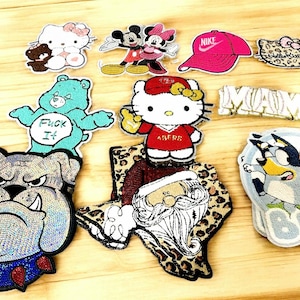 May include: A collection of iron-on patches featuring various cartoon characters, including a Care Bear, a bulldog, Santa Claus, Mickey Mouse, Hello Kitty, and a blue heeler dog. The patches are colourful and have a variety of designs.
