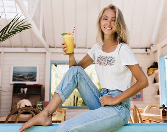 May include: A woman wearing a white t-shirt with the text "Flower Girl" and floral design sits on a blue bench, holding a yellow smoothie with a red and white striped straw. She is wearing blue jeans and has long blonde hair.