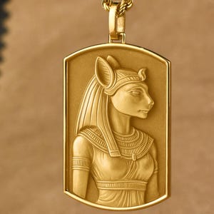 May include: A gold-colored pendant necklace featuring a rectangular pendant with a detailed relief of an Egyptian figure. The pendant is suspended from a matching gold chain. The figure has a cat-like head and is wearing a headdress.
