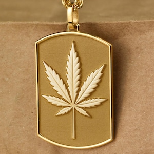 May include: Gold-colored pendant necklace featuring a cannabis leaf design. The pendant has a rectangular shape with a gold border and a detailed white leaf. The chain is a twisted rope style.