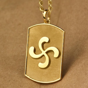 May include: A gold-colored pendant necklace featuring a rectangular tag with a stylized, curved symbol. The pendant hangs from a matching gold rope chain. The design is simple and elegant, with a brushed metal finish.