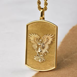 K&ouml;nnte beinhalten: Eine goldene Kettenkette mit einem goldenen Anh&auml;nger, der ein gepr&auml;gtes Bild eines Engels mit Fl&uuml;geln zeigt, der ein Schwert h&auml;lt und auf einer Schlange steht.