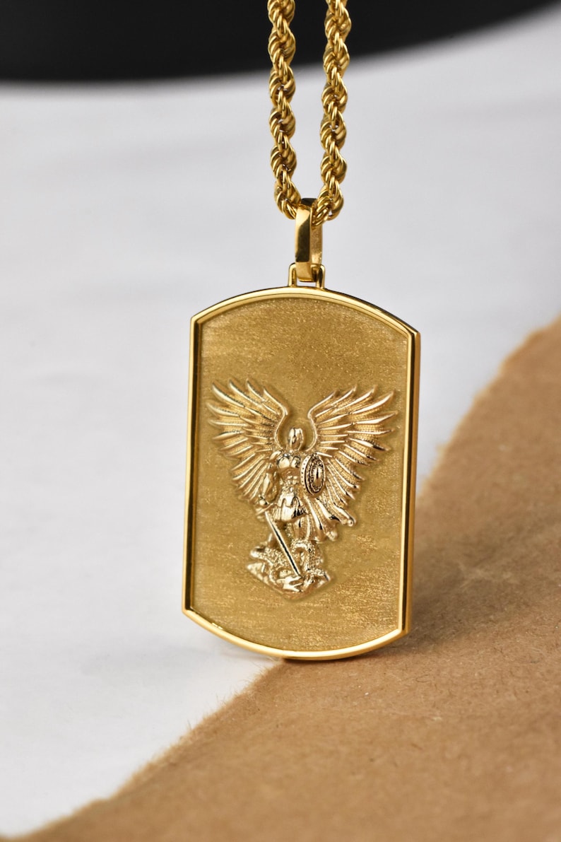 K&ouml;nnte beinhalten: Eine goldene Kettenkette mit einem goldenen Anh&auml;nger, der ein gepr&auml;gtes Bild eines Engels mit Fl&uuml;geln zeigt, der ein Schwert h&auml;lt und auf einer Schlange steht.