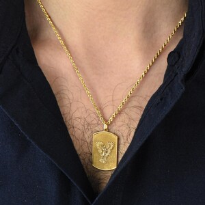 K&ouml;nnte beinhalten: Eine goldene Kettenkette mit einem goldenen Anh&auml;nger mit einem detaillierten Adler-Design. Der Anh&auml;nger ist rechteckig.