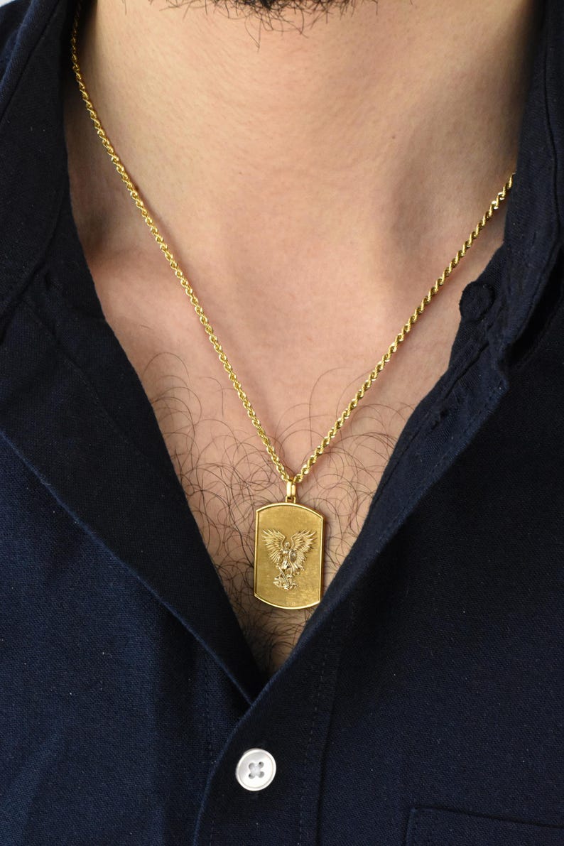 K&ouml;nnte beinhalten: Eine goldene Kettenkette mit einem goldenen Anh&auml;nger mit einem detaillierten Adler-Design. Der Anh&auml;nger ist rechteckig.