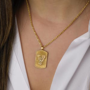 K&ouml;nnte beinhalten: Eine goldene Kettenkette mit einem goldenen Anh&auml;nger, der ein detailliertes Design eines Adlers mit ausgebreiteten Fl&uuml;geln zeigt. Der Anh&auml;nger ist in Form eines rechteckigen Hundemarken.