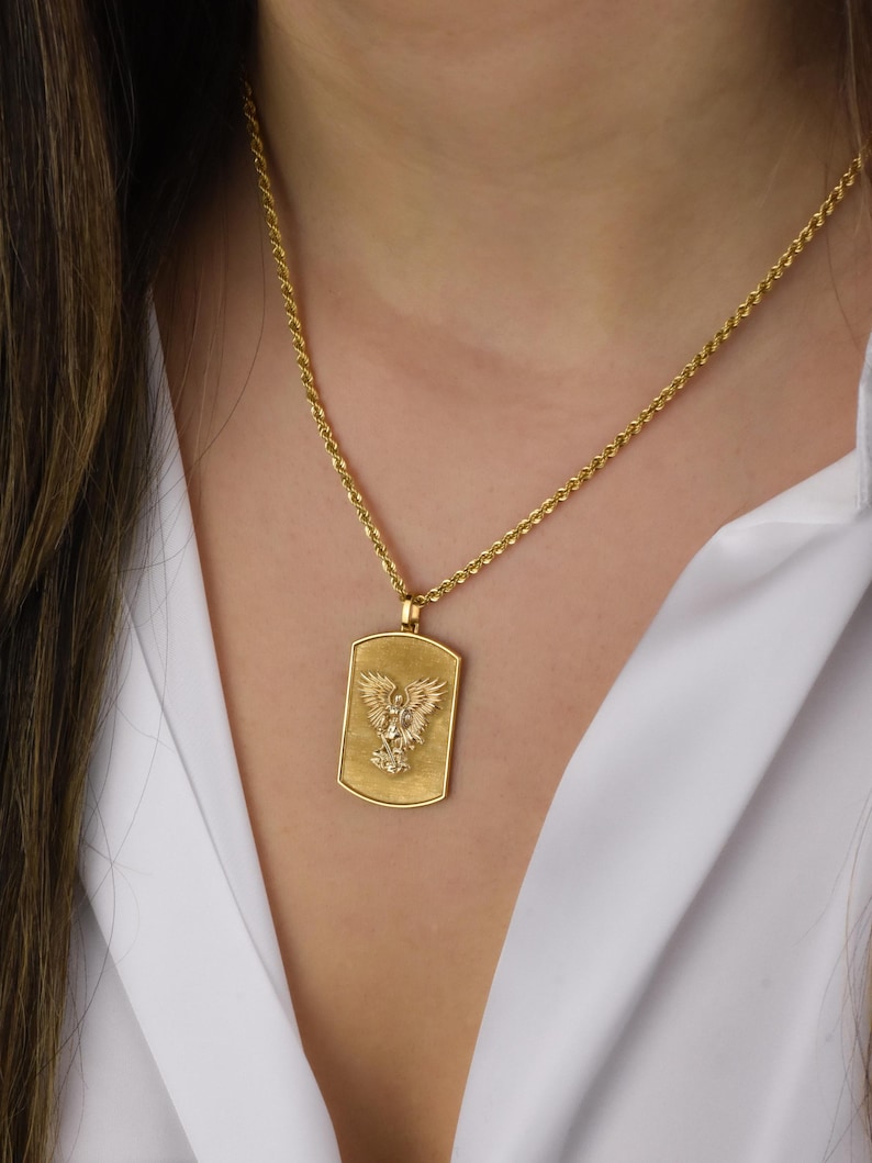 K&ouml;nnte beinhalten: Eine goldene Kettenkette mit einem goldenen Anh&auml;nger, der ein detailliertes Design eines Adlers mit ausgebreiteten Fl&uuml;geln zeigt. Der Anh&auml;nger ist in Form eines rechteckigen Hundemarken.