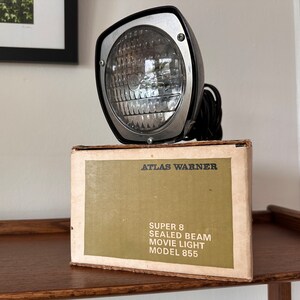 May include: A vintage Atlas Warner Super 8 Sealed Beam Movie Light, model 855, in its original box. The light is silver and black with a clear glass lens. The box is brown with green text.