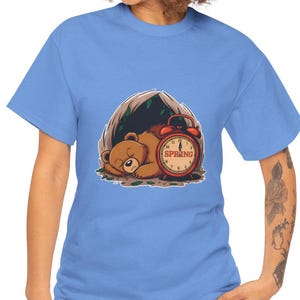 Könnte beinhalten: Ein blaues T-Shirt mit einer Cartoon-Illustration eines Bären, der unter einem Baum schläft, mit einem Wecker, auf dem "SPRING" steht.