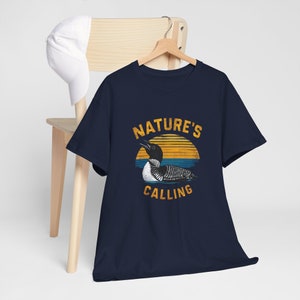 May include: Navy blue t-shirt featuring a vintage-style graphic of a loon in front of a sunset. The text "NATURE'S CALLING" is printed below the loon.