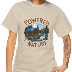 Op de afbeelding: Een beige t-shirt met een afbeelding van een hut in het bos met een windmolen en een waterval. De tekst "POWERED BY NATURE" is in een cirkel rond de afbeelding gedrukt.