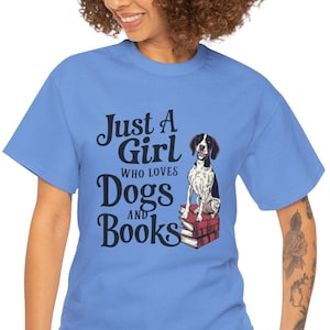 May include: A light blue t-shirt with the text "Just a Girl Who Loves Dogs and Books" in black. A black and white dog is sitting on a stack of red books.