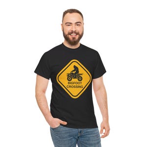 May include: Black t-shirt with a yellow warning sign graphic. The sign reads "Bigfoot Crossing" and depicts a Bigfoot driving an ATV.