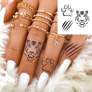 May include: Temporary tattoos featuring a tiger face, paw print, and claw marks in black ink. The tattoos are applied to fingers with white nail polish and gold rings.
