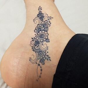 May include: A dark blue floral tattoo design on the side of a foot. The tattoo features a series of flowers, swirls, and dots. The design is intricate and detailed, with a focus on the natural curves of the foot.